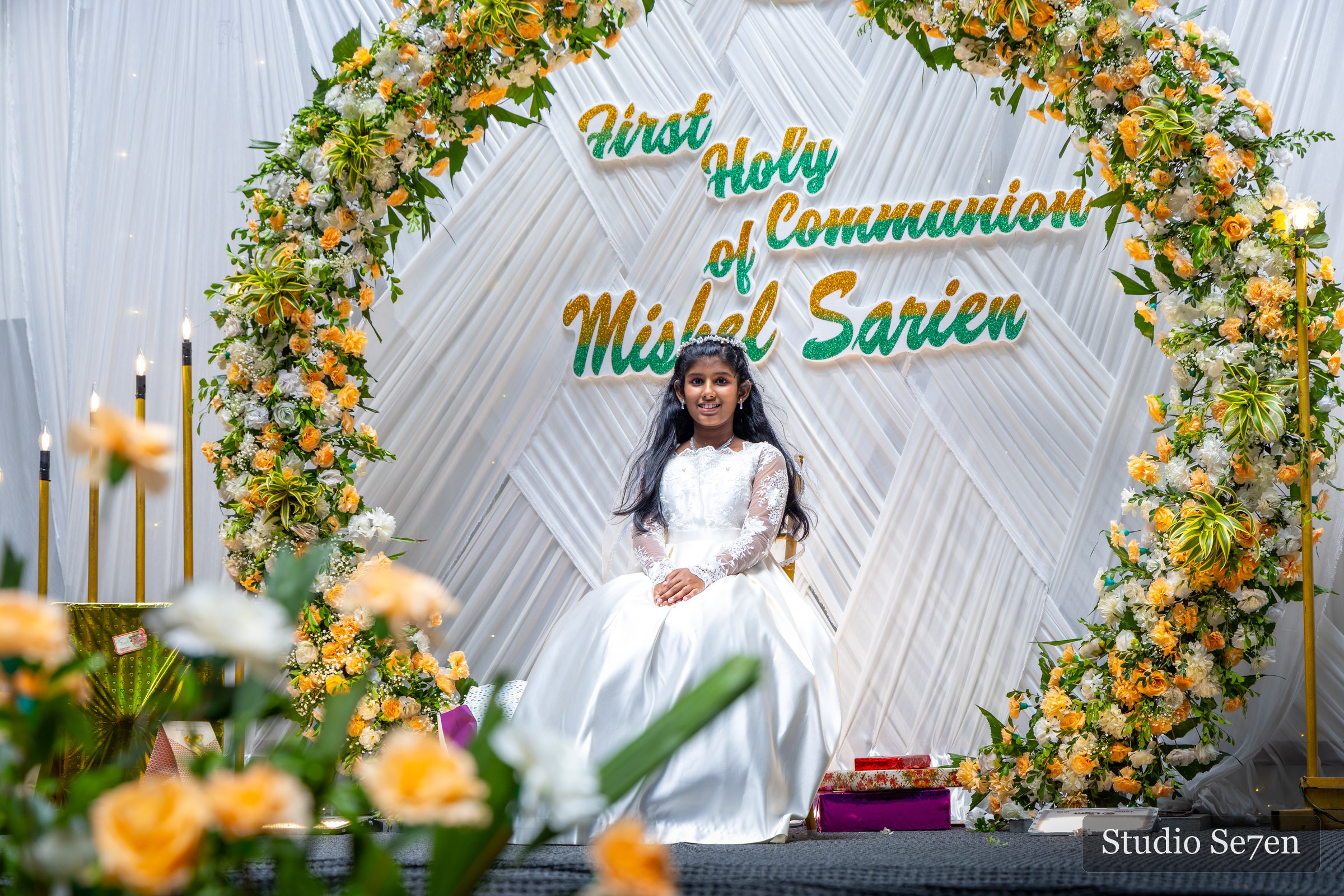 A child seated beneath a floral First Holy Communion backdrop