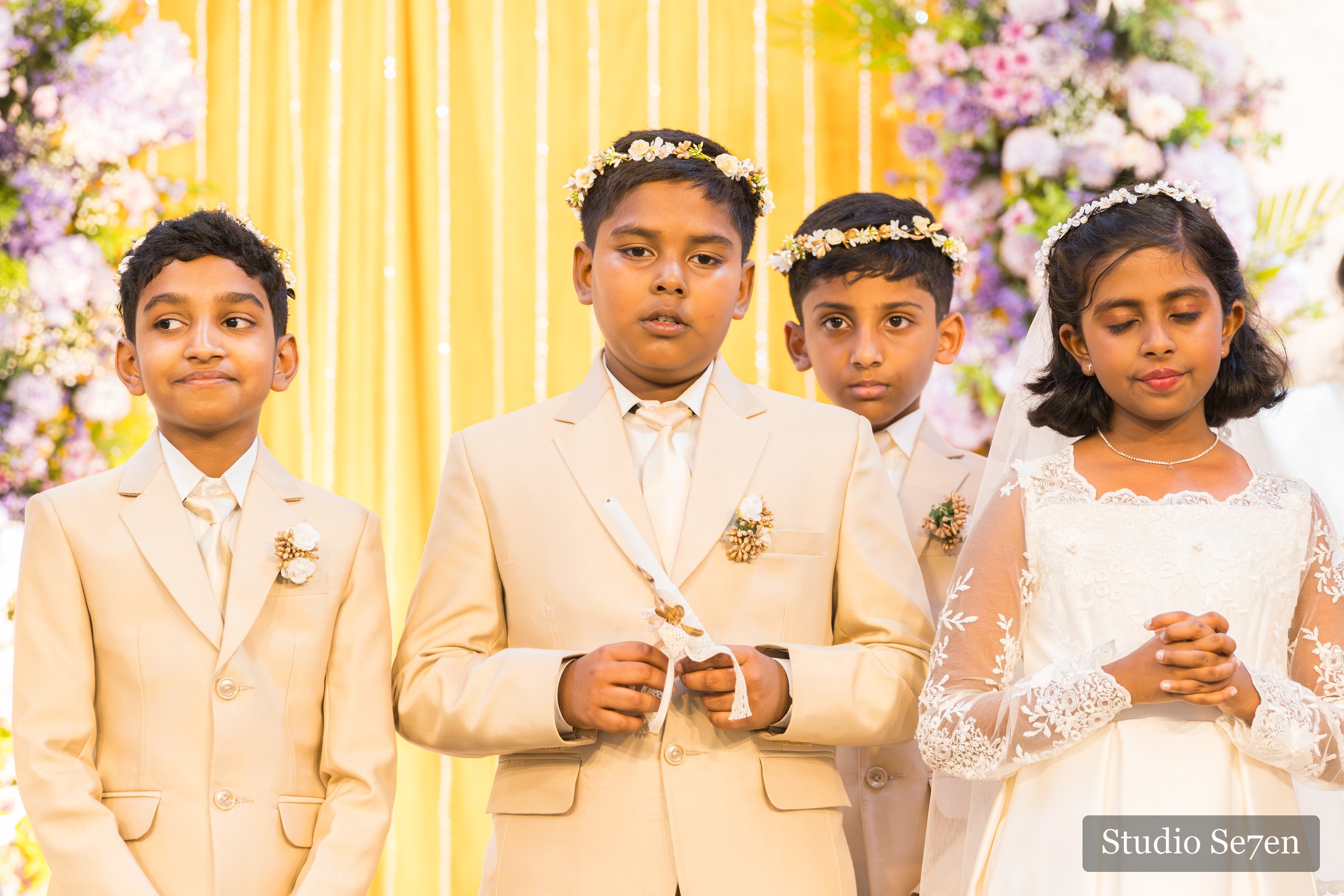 Four First Holy Communion children holding candles before a floral backdrop