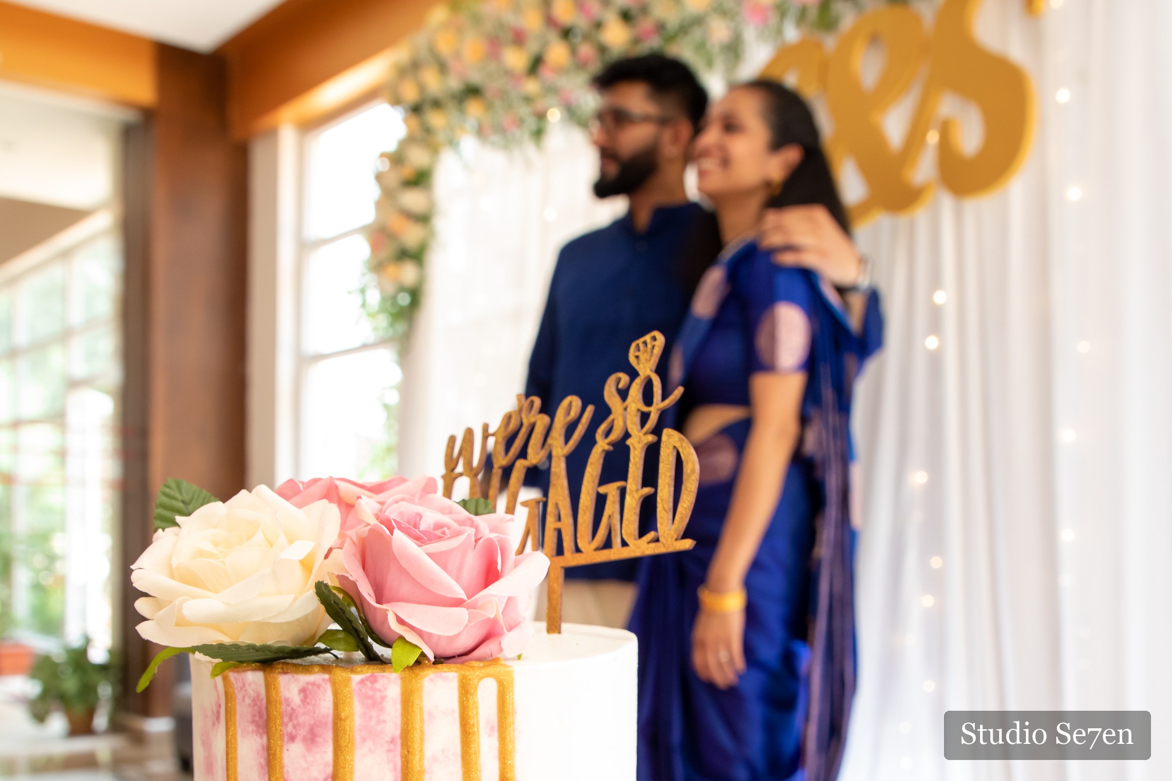 An engagement cake in the foreground with the couple sharing a quiet moment behind it