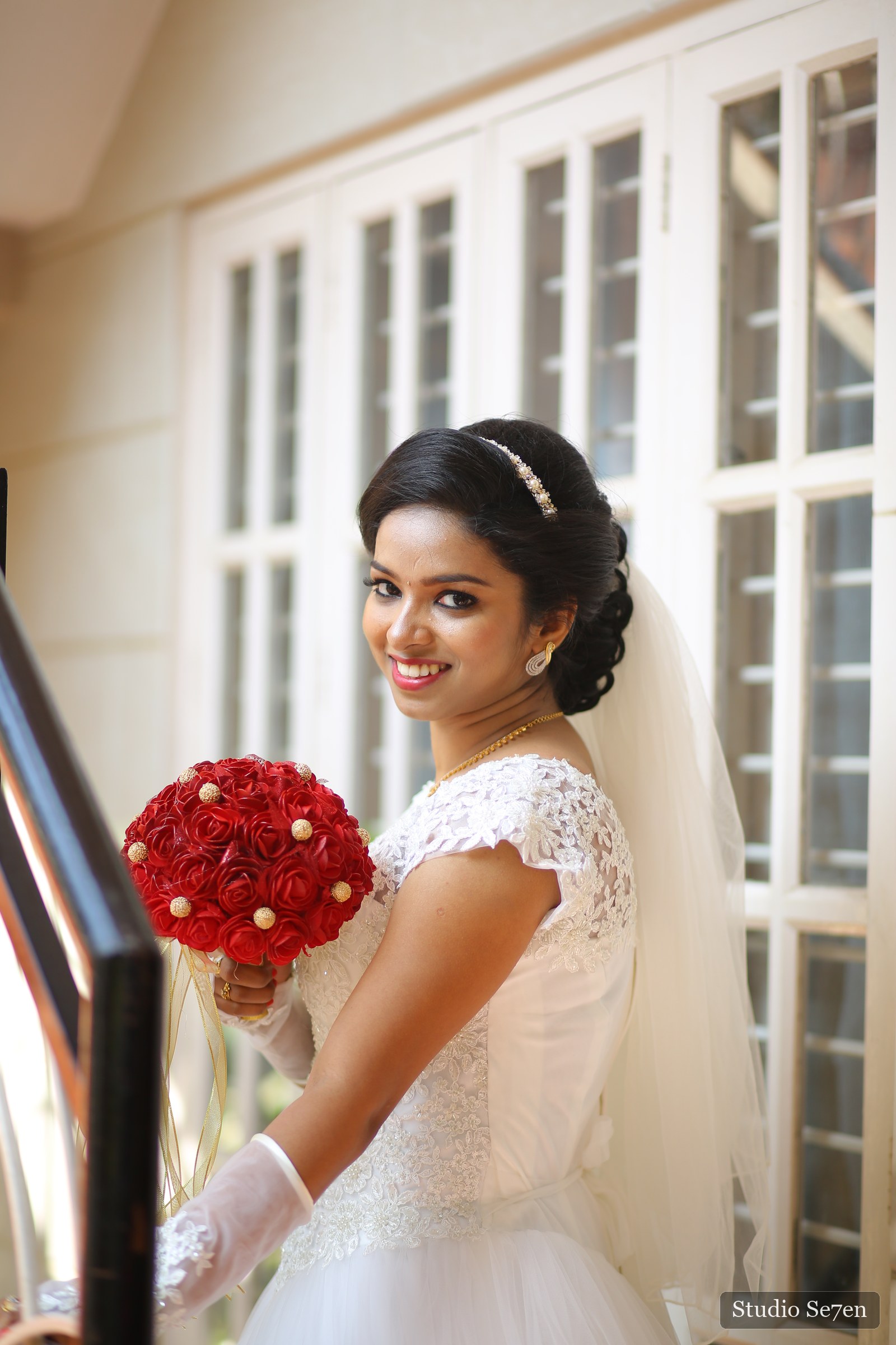 A bride holding a red rose bouquet beside a softly lit window